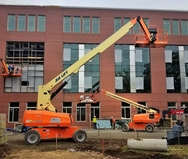 An orange and yellow JLG telescopic boom lift with its boom raised and basket against a brick building under construction, a second lift working behind it and workers on the ground.